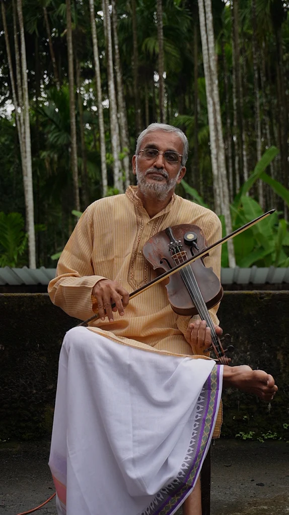 A teacher in a kurta and dhoti plays the violin outdoors among areca palms