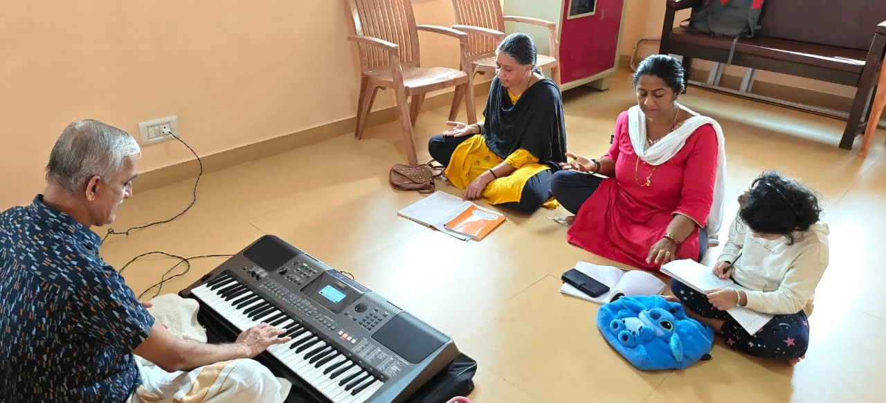 A teacher plays the keyboard while three students sit on the floor with their notebooks, learning a song