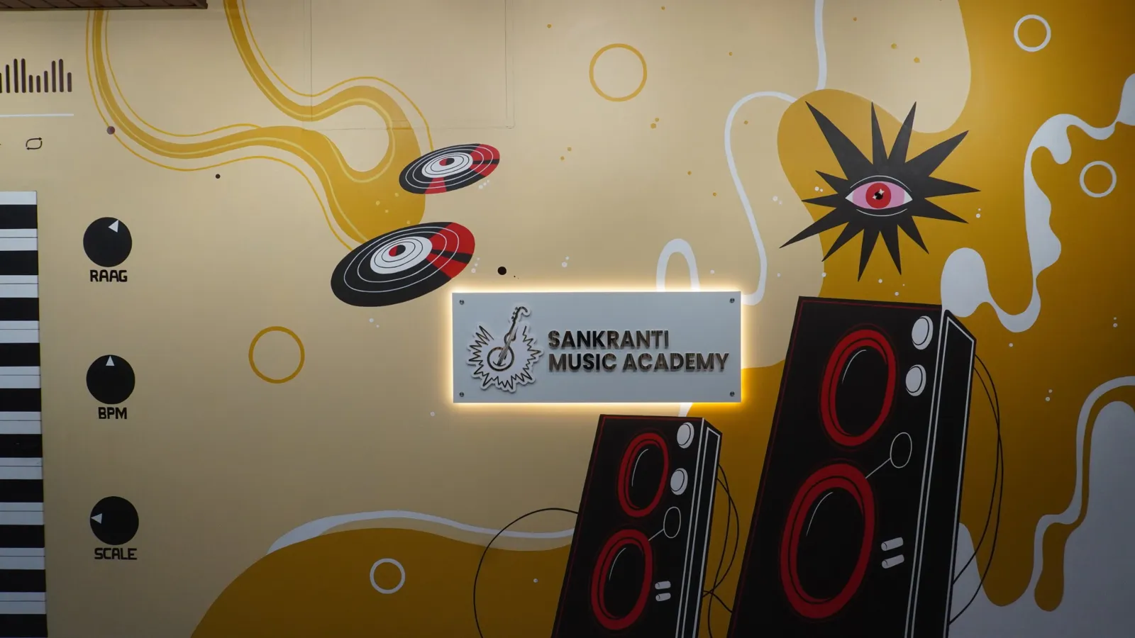 The painted mural wall at Sankranti Music Academy, with the lit academy sign between drawn speakers, records and keyboard keys