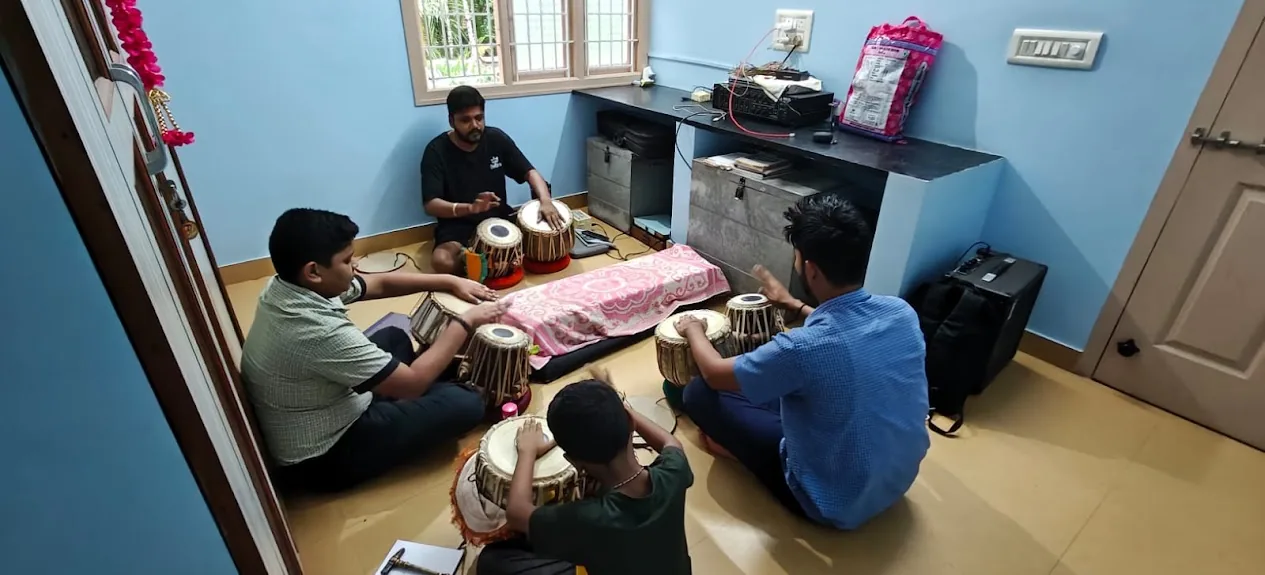 Three boys practise tabla on the floor with their teacher during a class