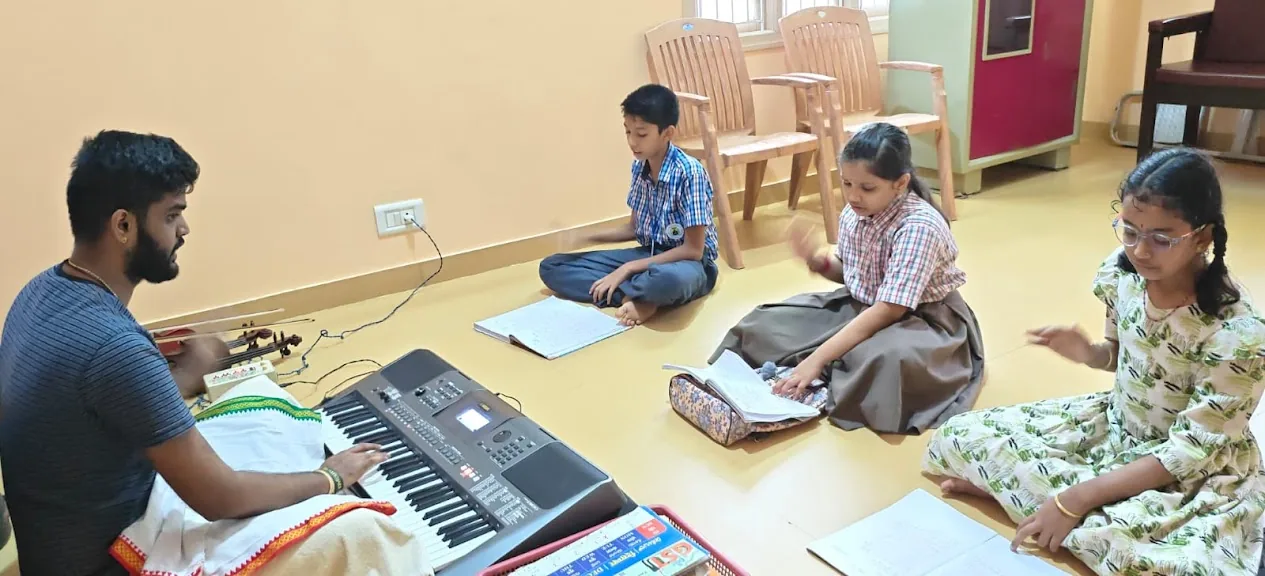 Three students on the floor keep the beat with their hands as their teacher plays the keyboard