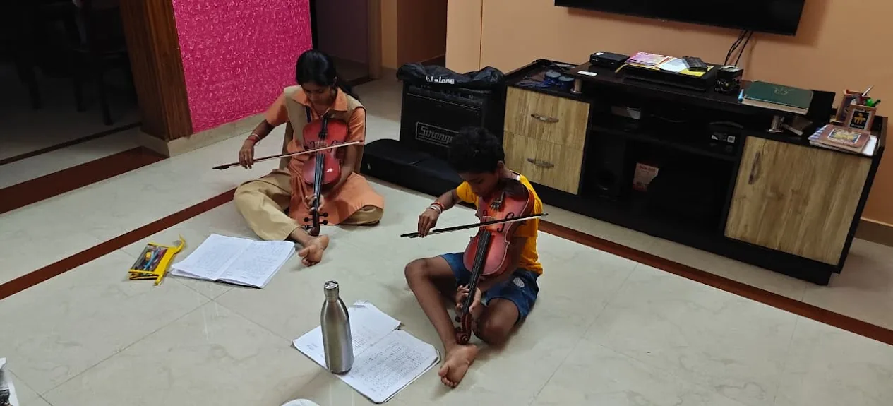 Two students sit on the floor playing the violin, with their notebooks open in front of them